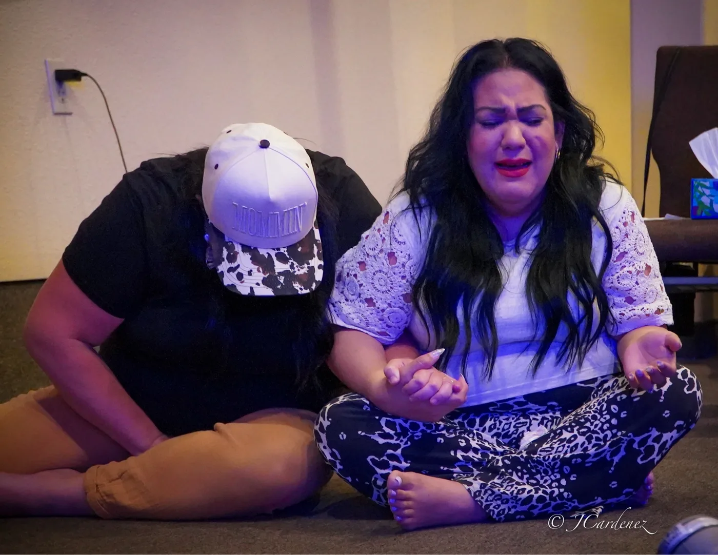 Two women seated on the floor in prayer, one with hands open and tears on her face